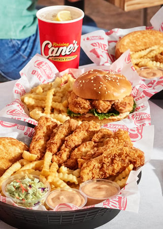 A table spread featuring multiple baskets of food, including chicken fingers, fries, coleslaw, and a chicken sandwich on a sesame seed bun, accompanied by a red drink cup with a lemon wedge.